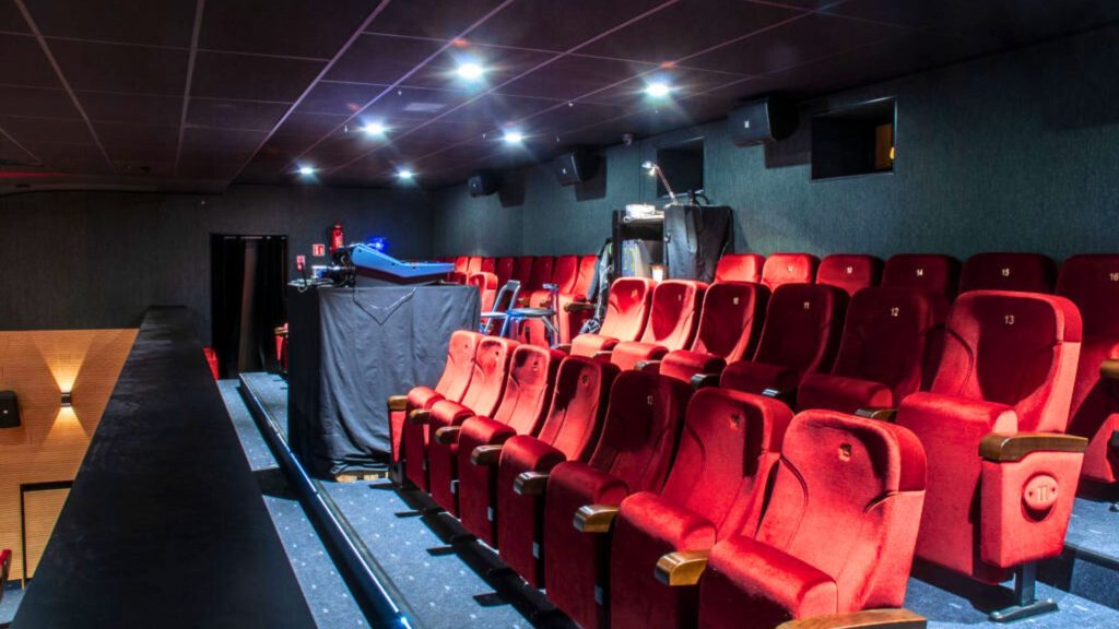 Zdjęcie przedstawia poziom balkonu sali widowiskowo-kinowej. Na pierwszym planie kilka rzędów czerwonych, tapicerowanych foteli z numerami na oparciach (widoczne numery od 11 do 16). Podłoga wyłożona ciemnoszarą wykładziną z niebieskim podświetleniem LED przy stopniach. Po lewej stronie balustrada balkonu z drewnianym wykończeniem i kinkietem. W centrum kadru profesjonalny projektor kinowy na statywie oraz stojak operatora przykryty czarną tkaniną. Na ścianie z tyłu zamontowane głośniki i monitory. Sufit ciemny z punktowymi reflektorami. Całość utrzymana w klimacie profesjonalnej sali kinowo-teatralnej.