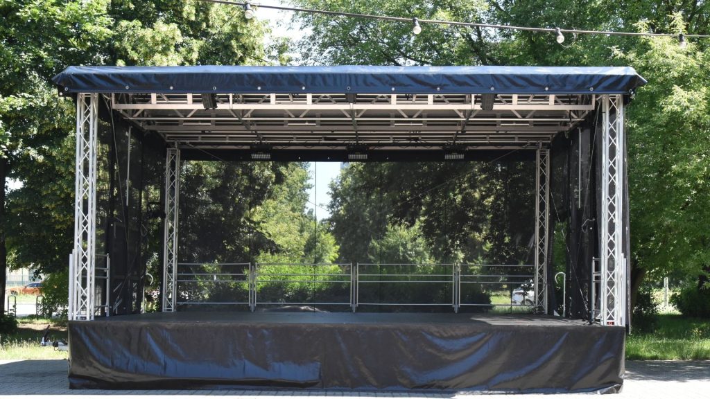 Mobilna scena plenerowa Domu Kultury „Świt" ustawiona w parku — scena estradowa na przyczepie z podwyższonym podestem obitym czarną tkaniną. Aluminiowa konstrukcja kratownicowa po bokach i nad sceną podtrzymuje zadaszenie kryte czarną plandeką. Na belkach sufitowych zamontowane reflektory. W głębi sceny przezroczyste panele tylnej ściany (backdrop), przez które widoczne są zielone drzewa. Na środku podestu metalowa barierka ochronna. W tle zielony park w słoneczny dzień.