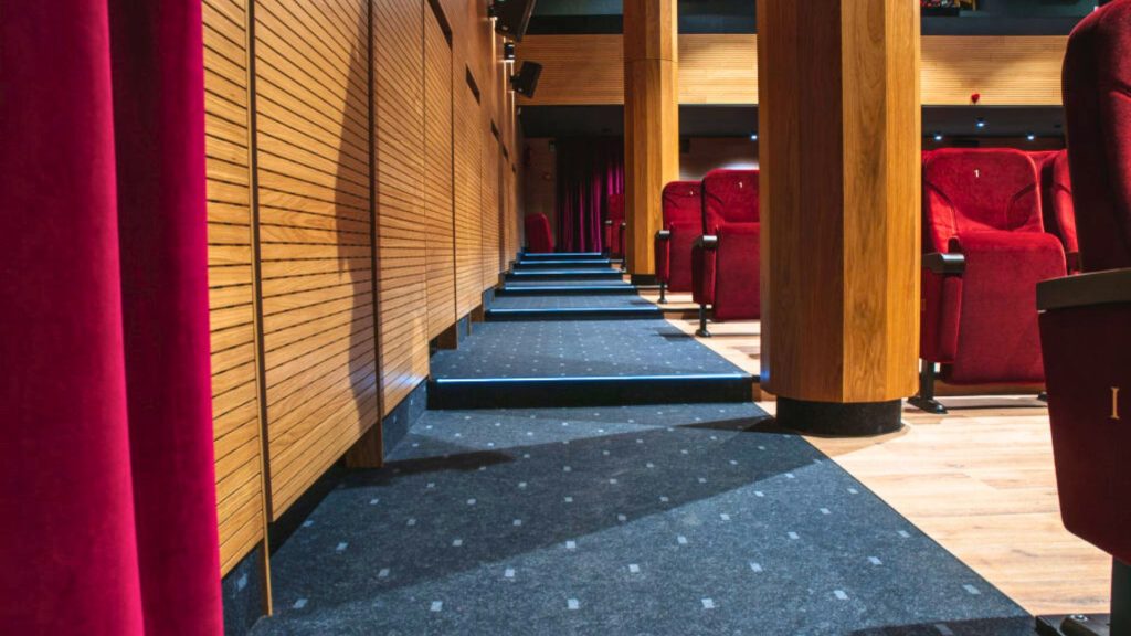Zdjęcie przedstawia boczny korytarz komunikacyjny na parterze sali widowiskowo-kinowej. Po lewej stronie ciemnoczerwona kurtyna sceniczna oraz ryflowana ściana z drewnianych lameli w ciepłym, jasnobrązowym odcieniu. W centrum kadru schody prowadzące ku górze widowni — stopnie wyłożone ciemnoszarą wykładziną wzorzystą, z niebieskim podświetleniem LED wbudowanym w każdy stopień. Po prawej stronie czerwone, tapicerowane fotele teatralne z numerami rzędów. Całość utrzymana w eleganckiej stylistyce — drewno, czerwień i stalowoszara wykładzina tworzą spójną, teatralną aranżację wnętrza.