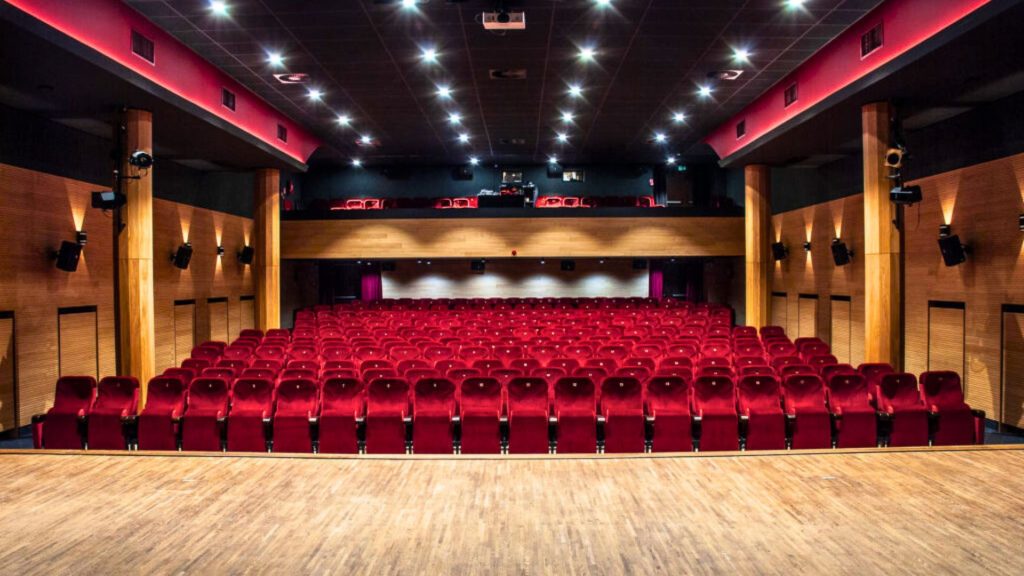 Na pierwszym planie jasna, drewniana podłoga sceny. W centrum kadru kilkanaście rzędów czerwonych, tapicerowanych foteli teatralnych ustawionych amfiteatralnie. Po bokach sali drewniane ściany z kolumnami i kinkietami emitującymi ciepłe, pomarańczowe światło. Sufit ciemny z białymi punktowymi reflektorami scenicznymi oraz różowo-czerwonym podświetleniem obwodowym. W głębi widoczny balkon z dodatkowym rzędem siedzeń oraz kabina techniczna (reżyserka).